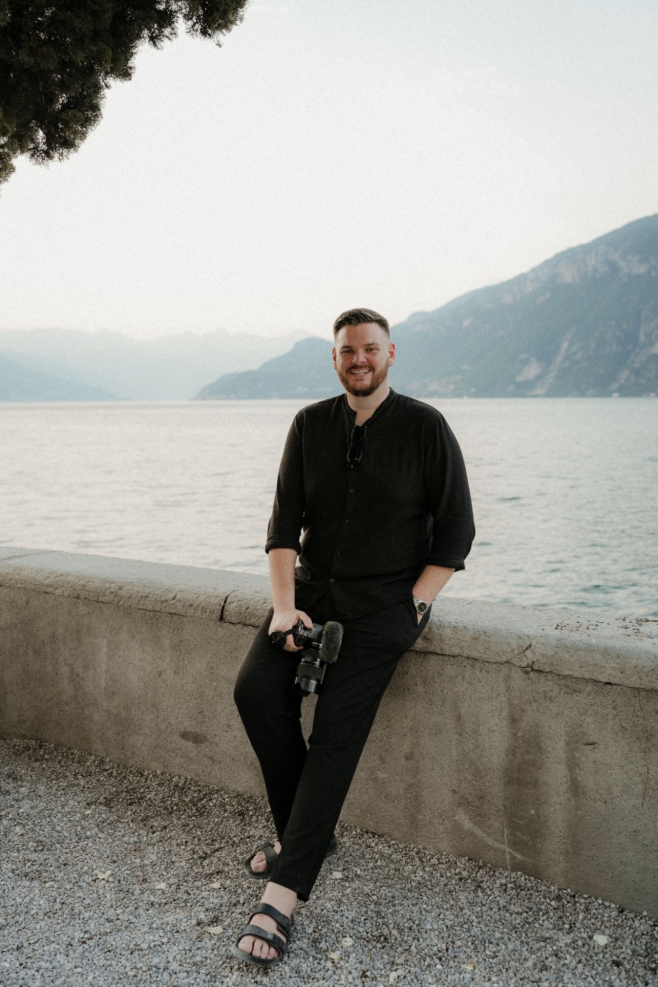 Matt from PS Visuals with his camera, sitting on a lakeside wall with mountains behind